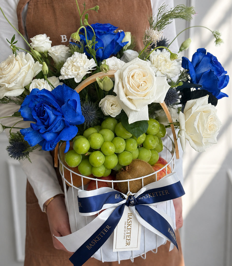 Luxury gift basket with blue roses, white carnations, green grapes, and fruits, tied with navy and white ribbons.