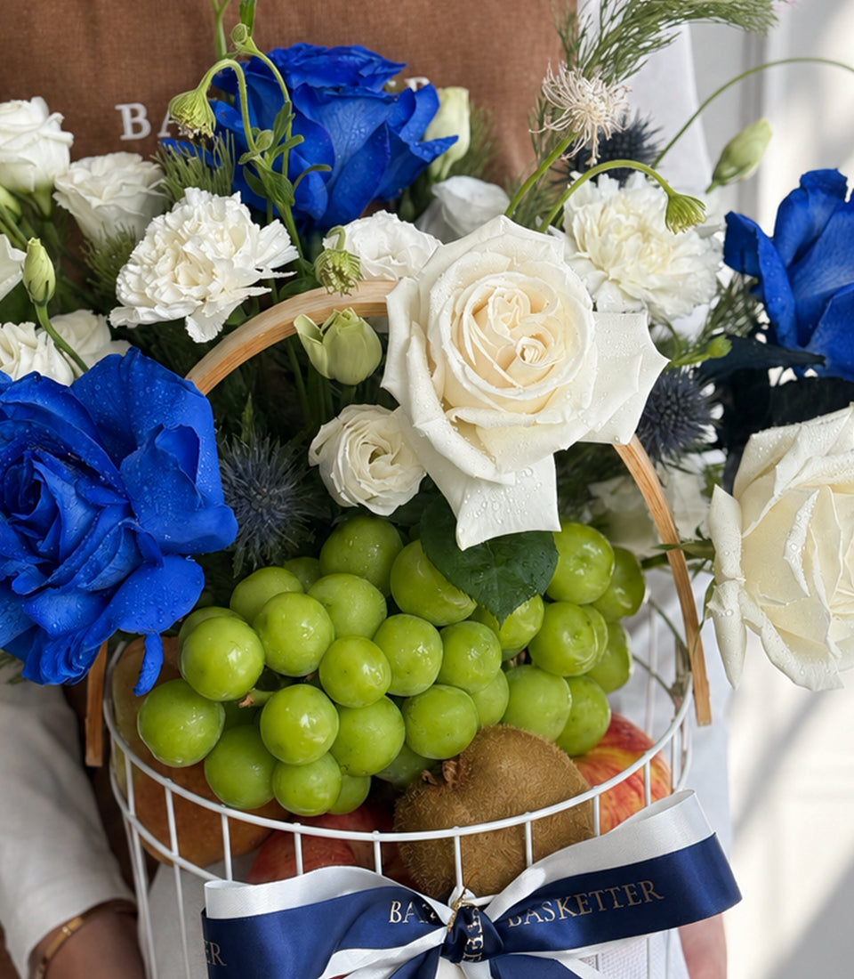 Luxury floral gift basket with blue roses, white carnations, green grapes, kiwis, and apples, tied with navy ribbon.