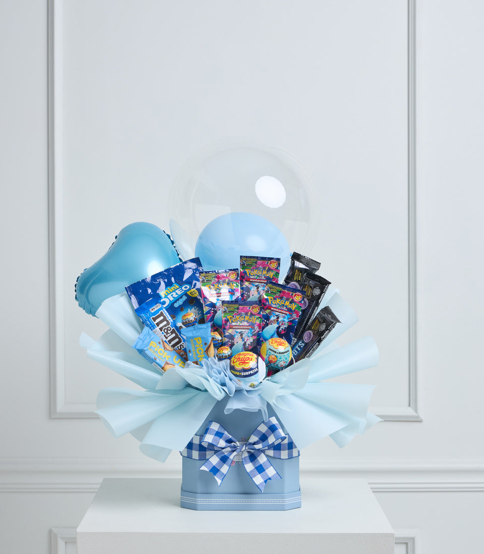 Blue gift bouquet with balloons, Pokemon cards, snacks and candy in light blue wrapper with blue gingham bow on white pedestal.