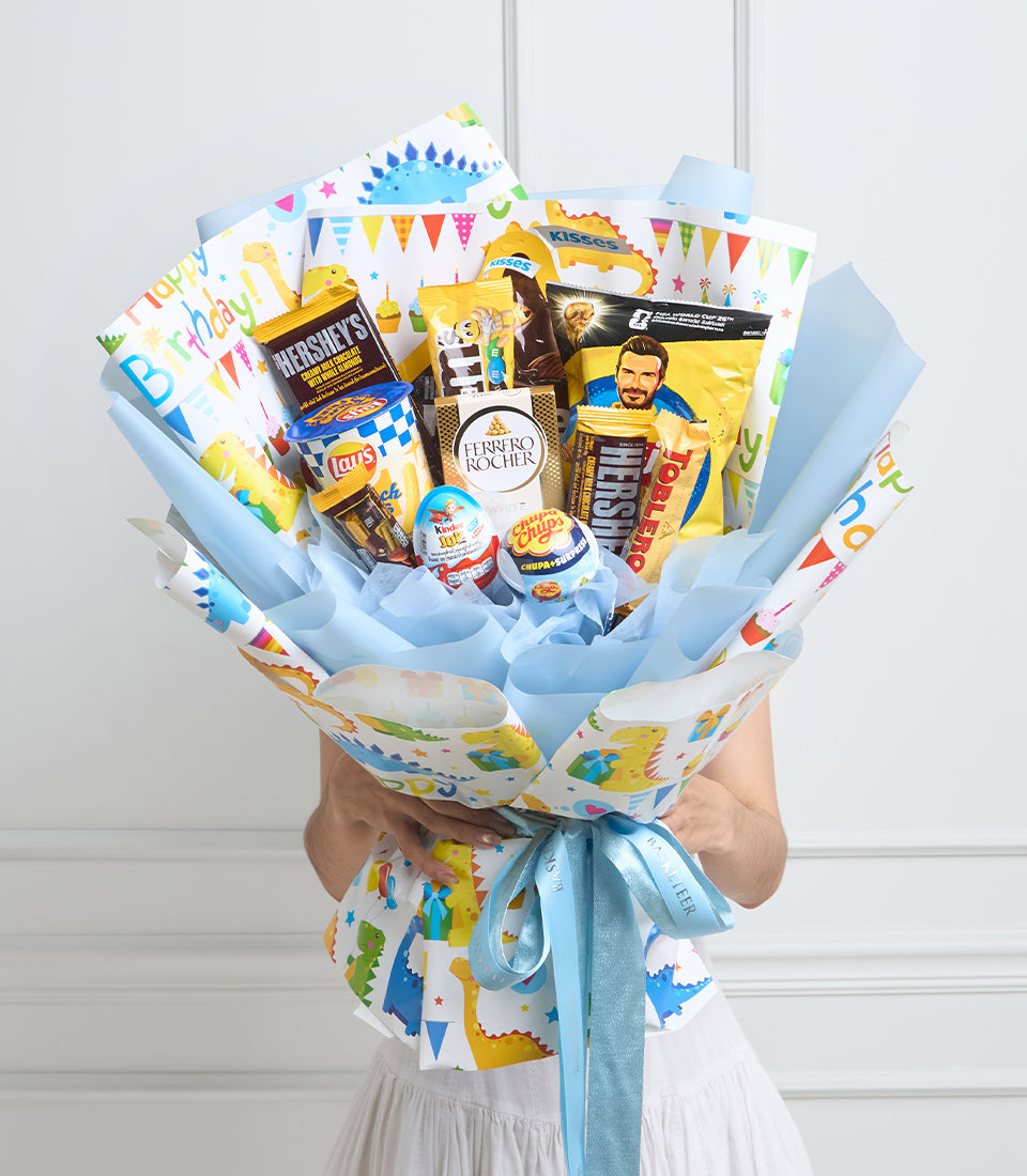 # Alt Text

Colorful birthday gift bouquet wrapped in blue and white paper with dinosaur and party designs, filled with snacks, chocolates, and candies, tied with light blue ribbon.