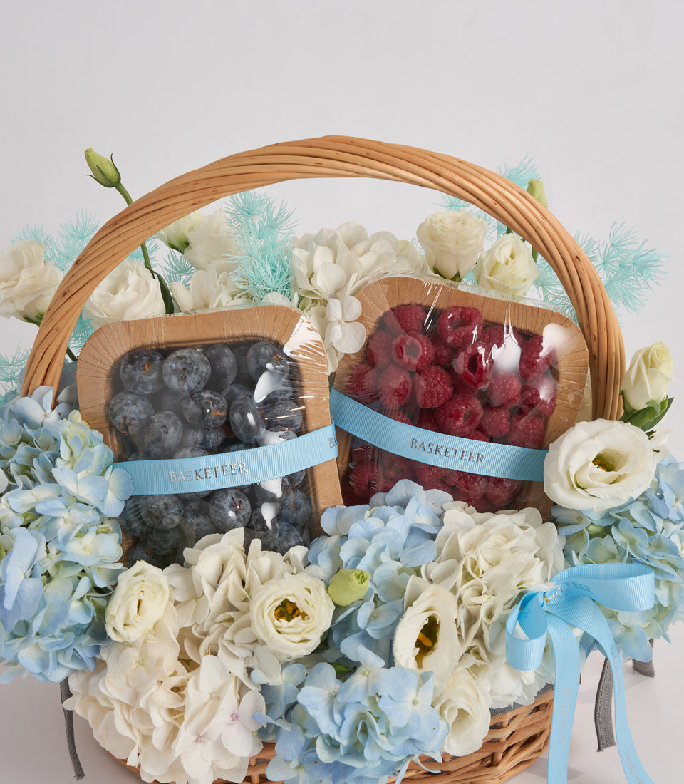 Wicker basket with fresh berries in clear containers, white roses, blue hydrangeas, and decorative blue ribbons on neutral background.