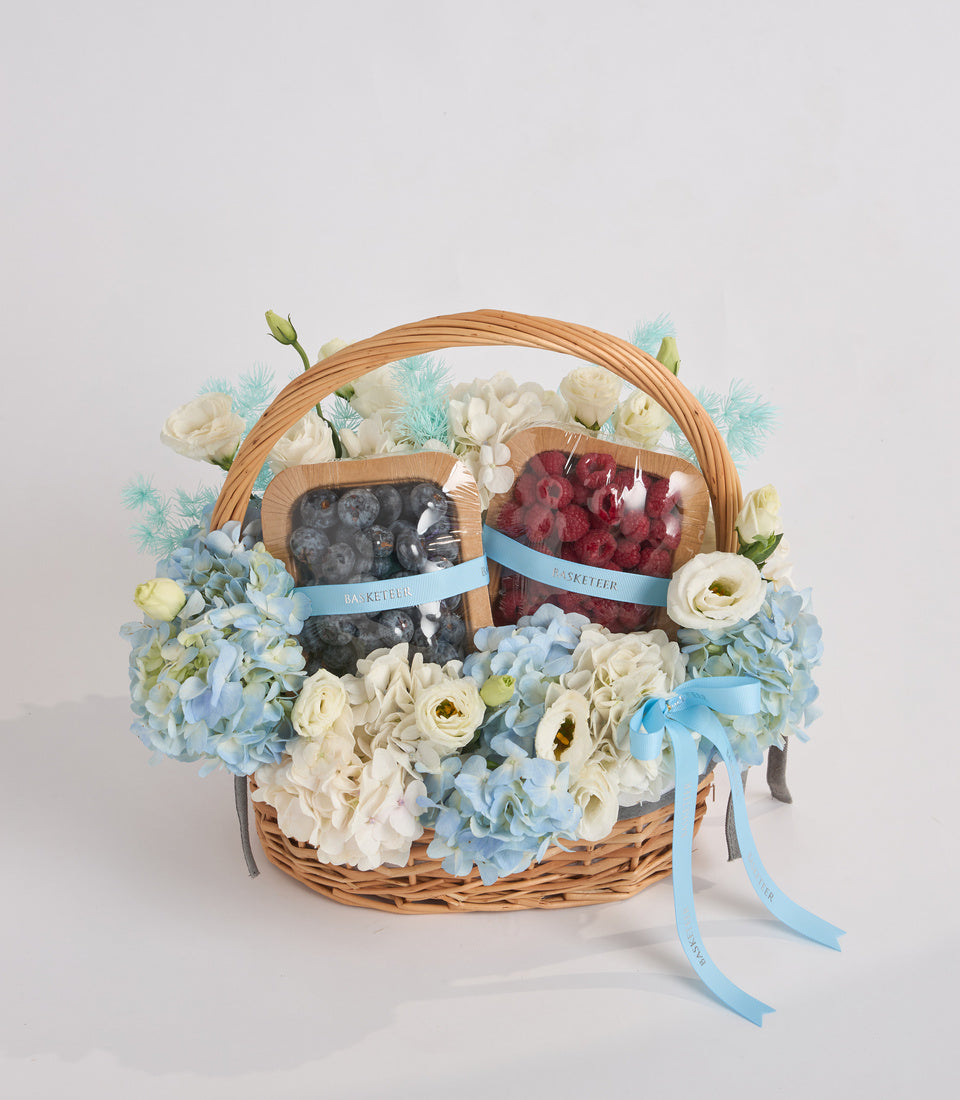 Wicker basket with blue and white flowers, blueberries and raspberries in containers with blue ribbons, decorated with cream roses and hydrangeas.