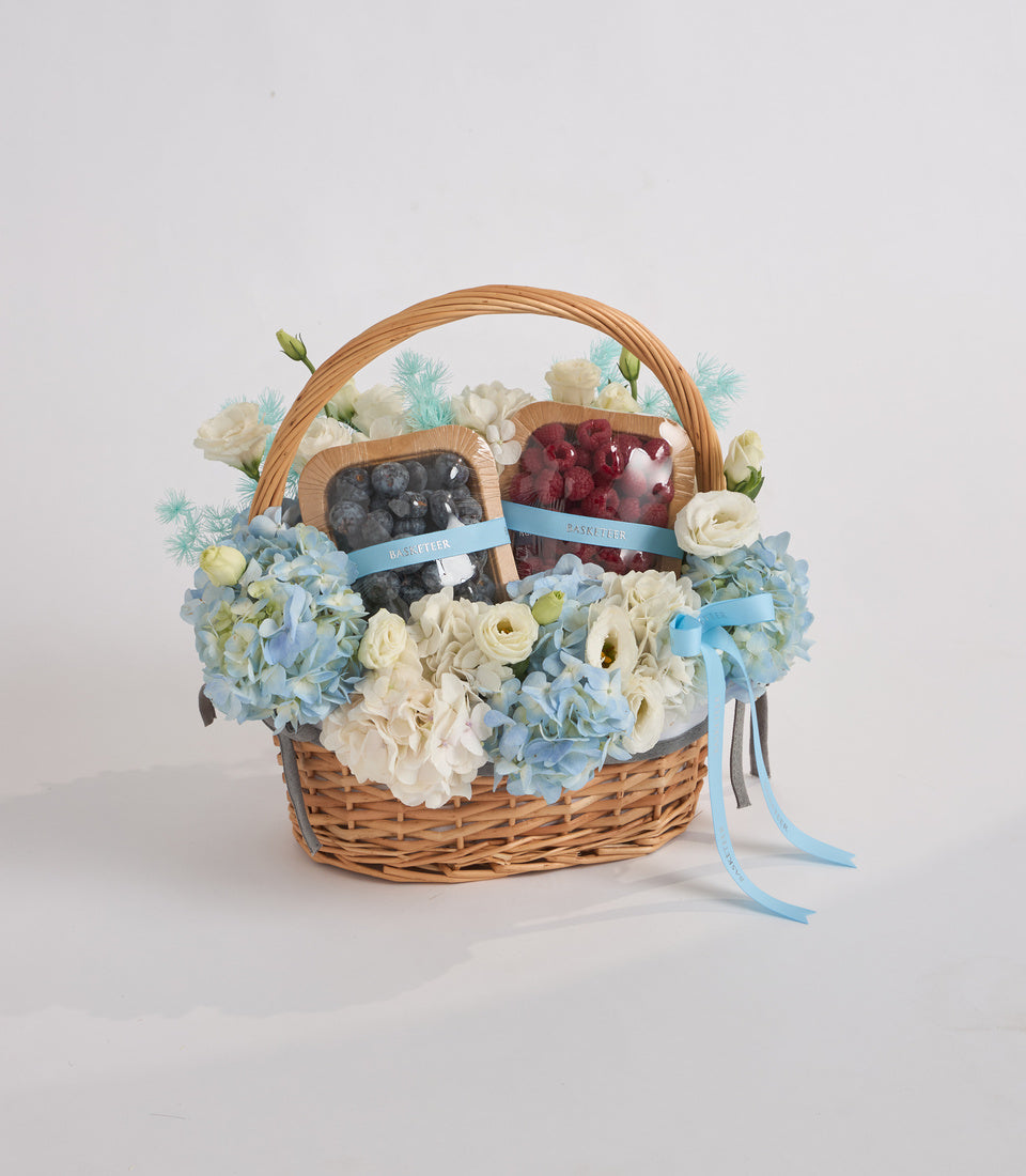 Wicker basket filled with blue hydrangeas, white roses, and turquoise filler flowers, topped with blueberry and raspberry containers tied with blue ribbons.