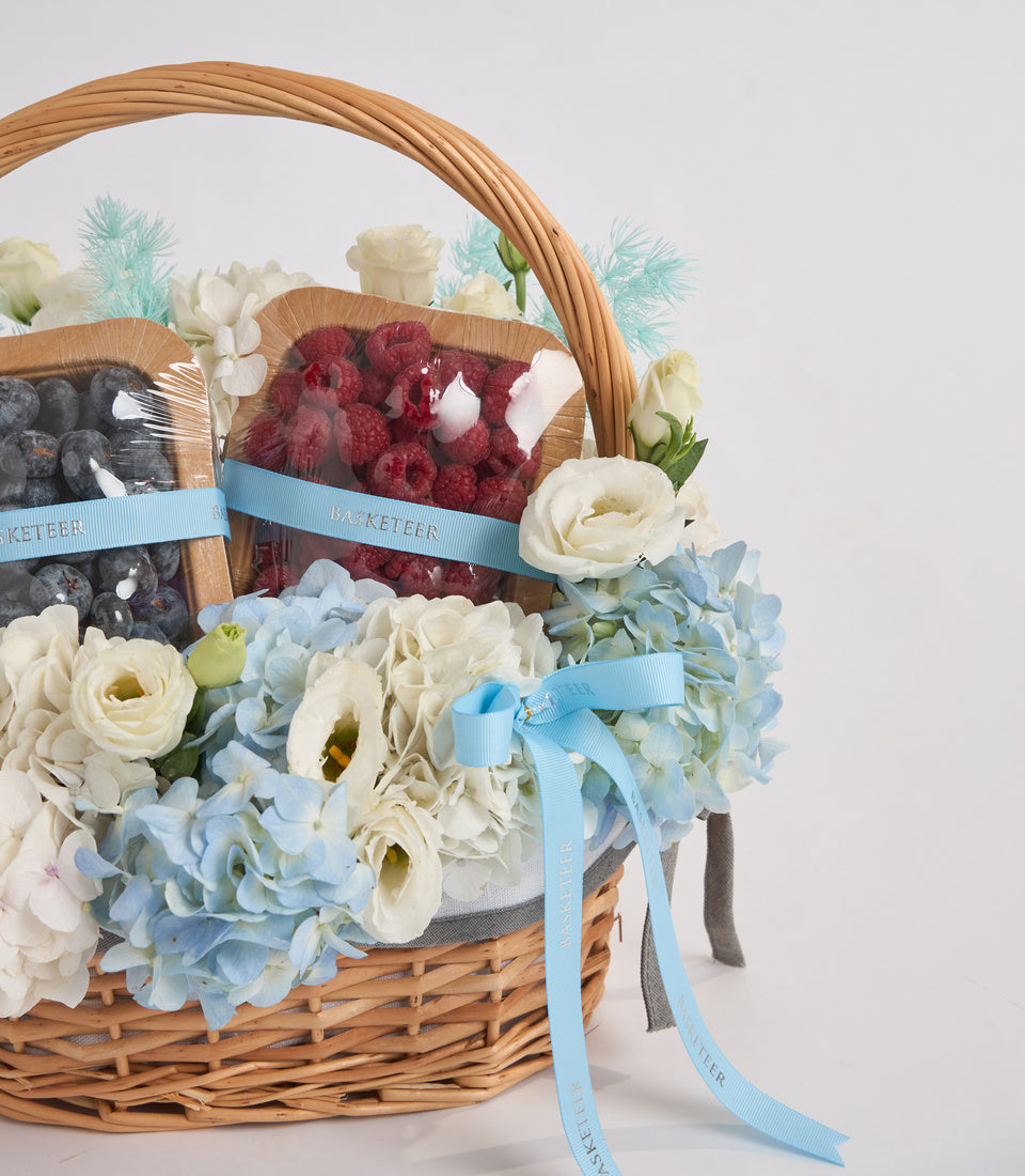 Wicker basket filled with white roses, blue hydrangeas, raspberries, blueberries, and turquoise ribbon bows against a white background.