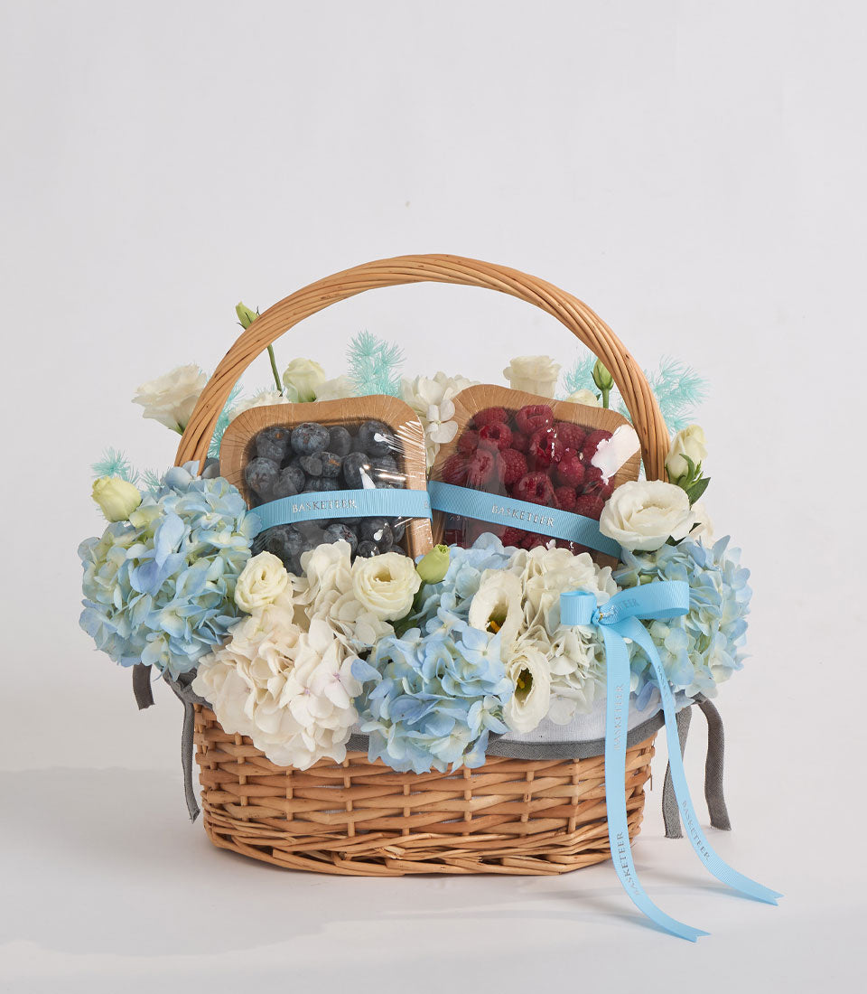 Wicker basket with cream and blue hydrangeas, white roses, filled with blueberries and raspberries, blue ribbons, elegant gift arrangement.