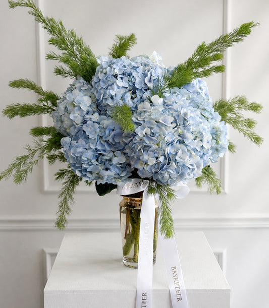 Luxurious pastel blue hydrangea arrangement with lush green foliage in a clear glass vase