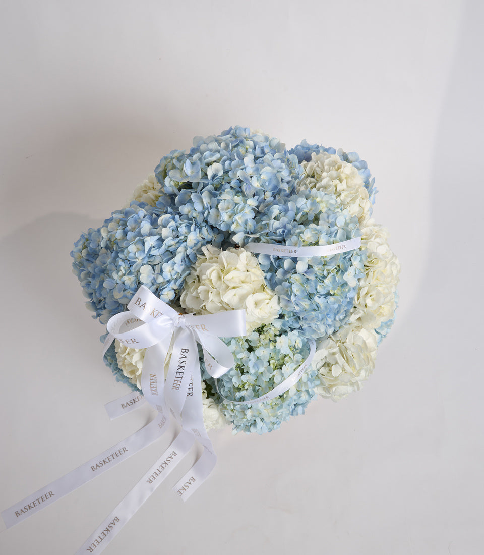 Luxurious floral arrangement with blue and white hydrangeas tied with white silk ribbon branded "Basketeer" against neutral background.