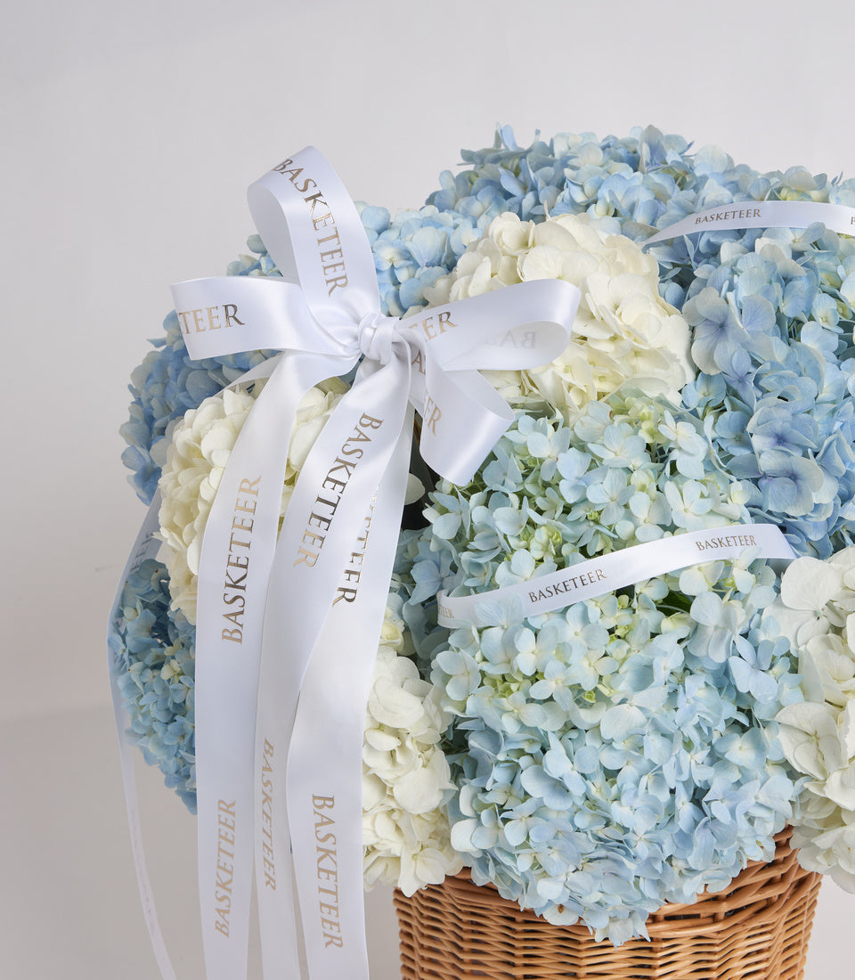 Elegant floral gift basket with blue and white hydrangeas in woven wicker, adorned with white "Basketeer" branded ribbons and bow.