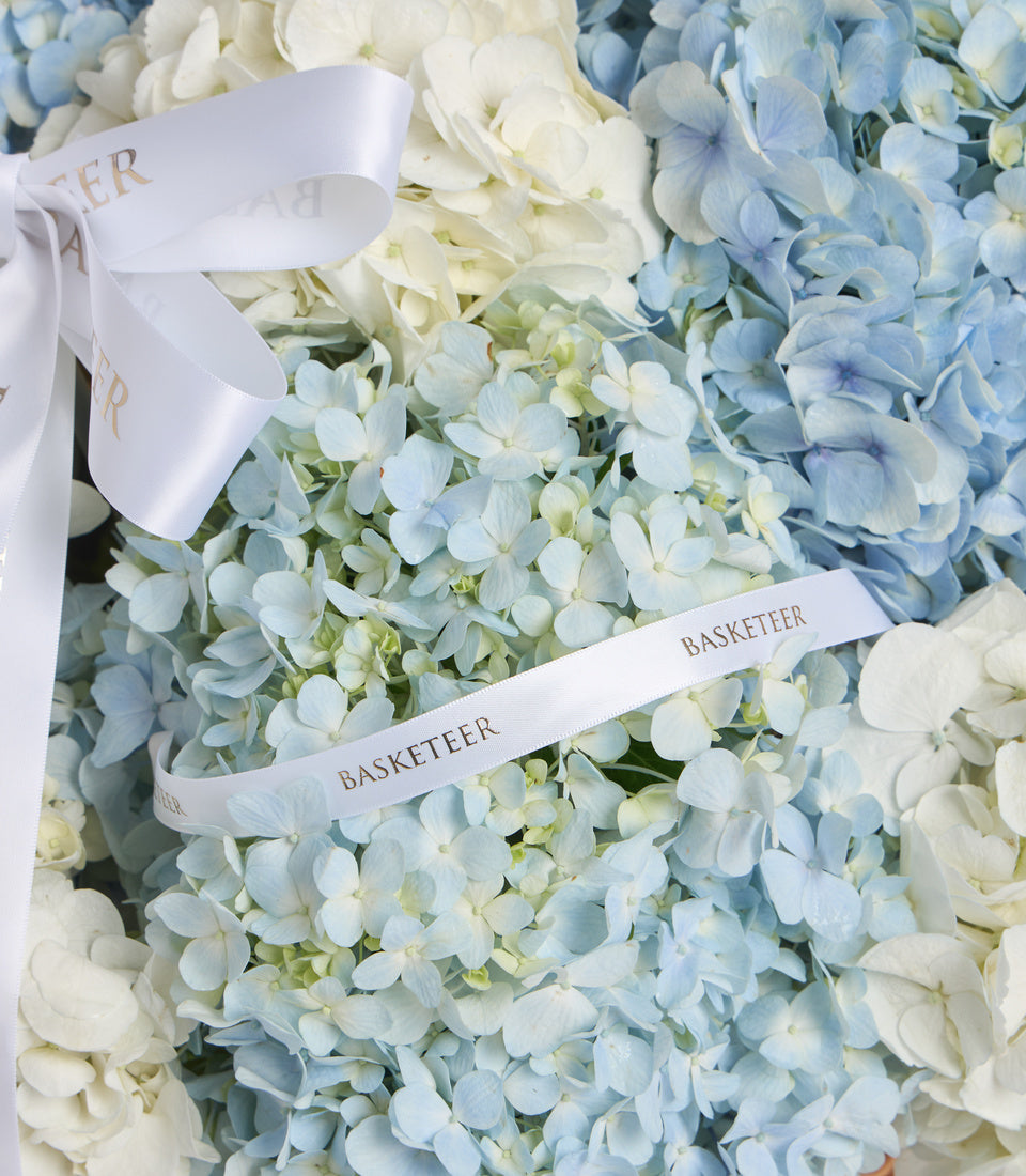 White silk ribbons with "BASKETEER" text tied around a lush bouquet of blue and cream hydrangeas in soft, romantic style.