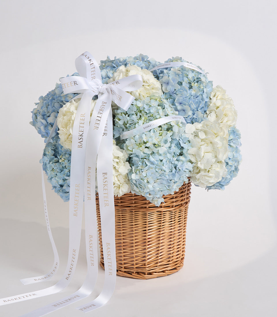 Elegant floral arrangement in wicker basket with blue and white hydrangeas, topped with white "Basketeer" ribbon bow against neutral background.