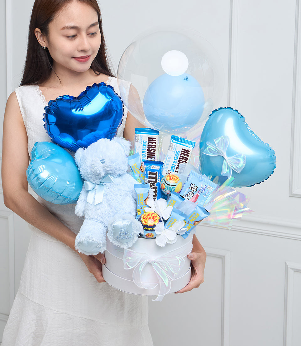 Woman holding blue and white gift arrangement with foil balloons, plush teddy bear, candies, and chocolate bars in white box.