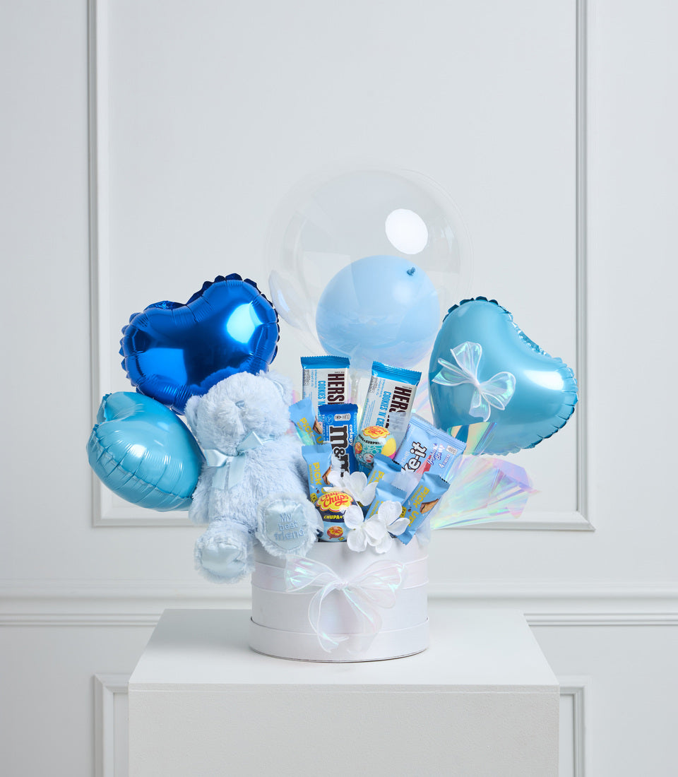 Blue foil balloons, white teddy bear, and assorted snacks arranged in a white box with ribbon bow against a light background.