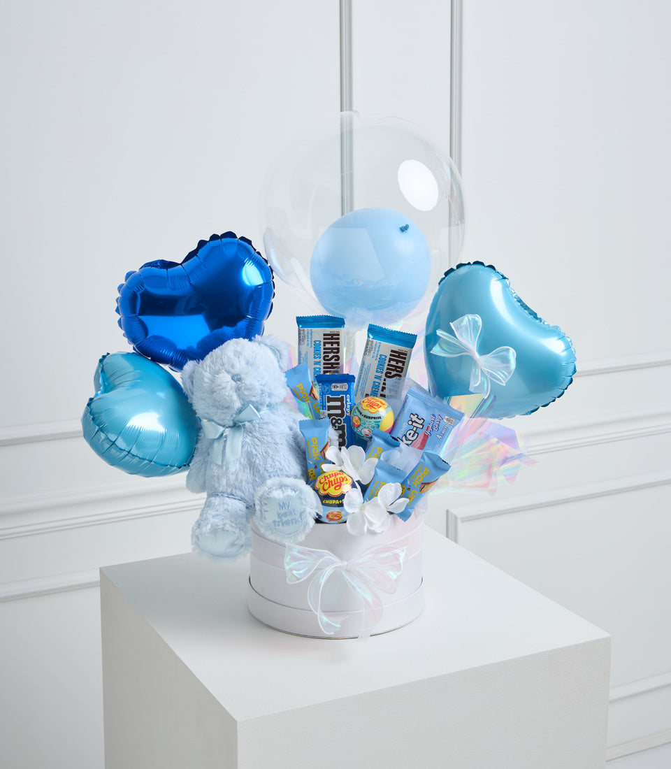 Blue foil balloons, white teddy bear, chocolate bars, and white flowers in a white box with iridescent ribbon bow.