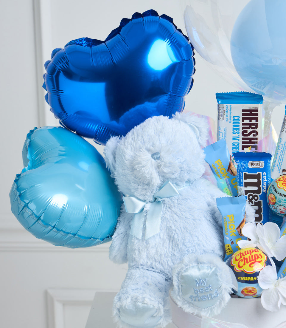 White plush teddy bear holding blue foil balloons, surrounded by candy and snacks including chocolate bars and lollipops.