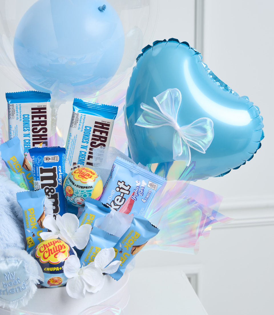 Blue heart-shaped foil balloon with white bow surrounded by candy and chocolate treats including Hershey's, M&Ms, and Chupa Chups lollipops, with decorative white flowers and balloons.