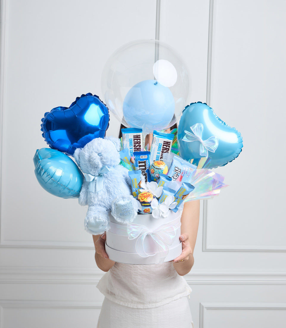 Blue and white gift arrangement with balloons, stuffed animal, chocolates, and candy in a white box with ribbon bow.