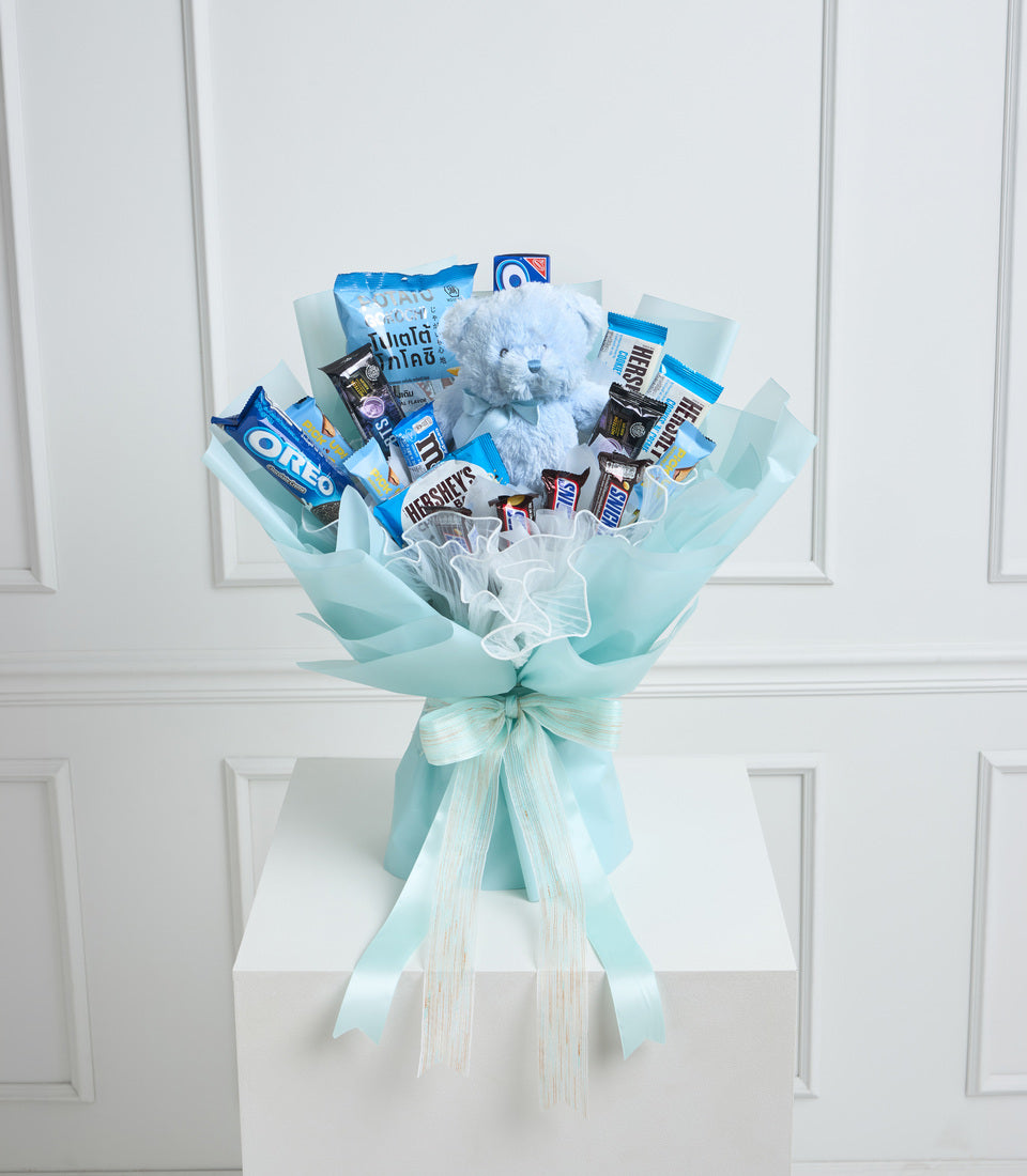 Light blue wrapped bouquet with snacks, candies, Oreo cookies, Hershey's chocolate, and a plush blue teddy bear on white background.