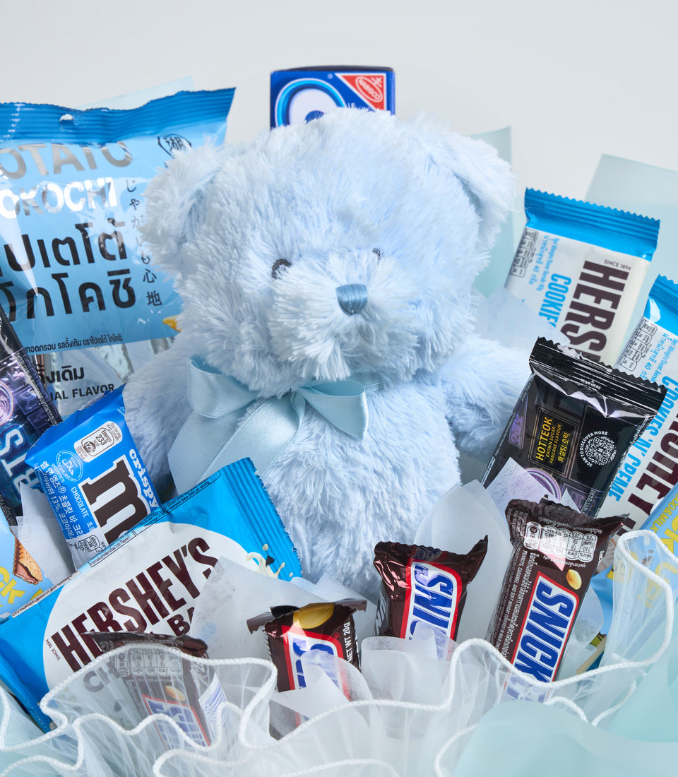 White fluffy teddy bear with blue ribbon bow surrounded by assorted chocolate and candy treats in blue and brown packaging.