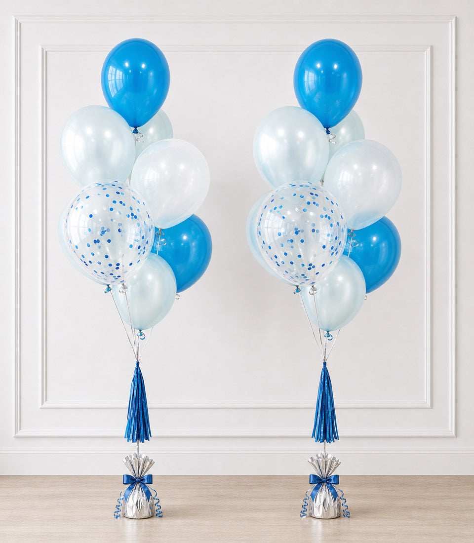 Two blue and white balloon bouquets with confetti-filled balloons, blue tassels, and weighted silver bases against a white paneled wall.