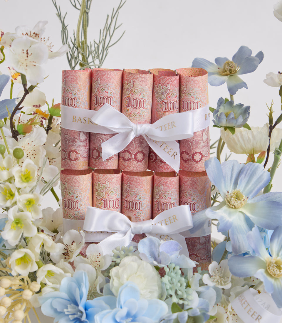 Stack of pink £100 banknote-patterned rolls tied with white satin ribbons, surrounded by white and blue flowers.
