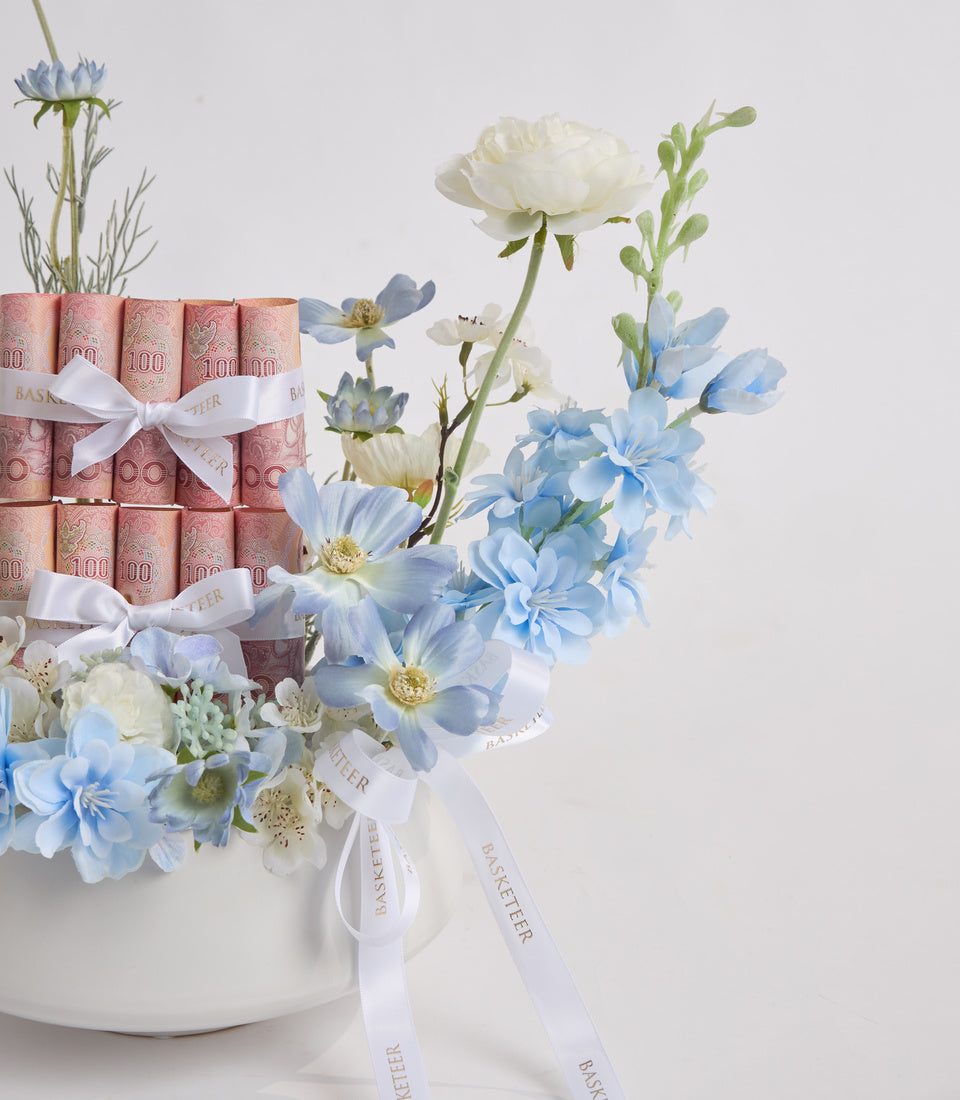 Tiered cake with stacked banknotes, white ribbons, and fresh flowers in blue and white tones in a white ceramic base.
