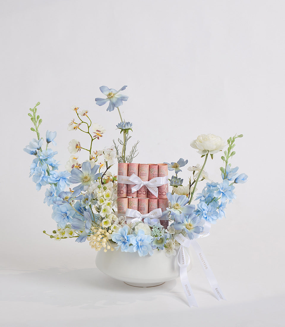 Luxury floral arrangement with pink rolled cosmetics, blue and white flowers, cream roses in white ceramic bowl with white ribbon bows.