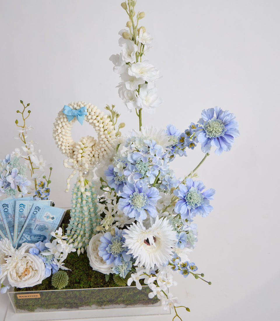 Luxury floral arrangement with blue and white flowers, pearl beaded garland, moss base, and decorative euro bills in clear acrylic box.