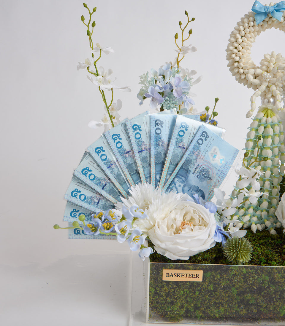 Luxury floral arrangement featuring white roses, blue flowers, and orchids with fanned £60 banknotes, pearl beads, and moss-lined container with blue ribbon.