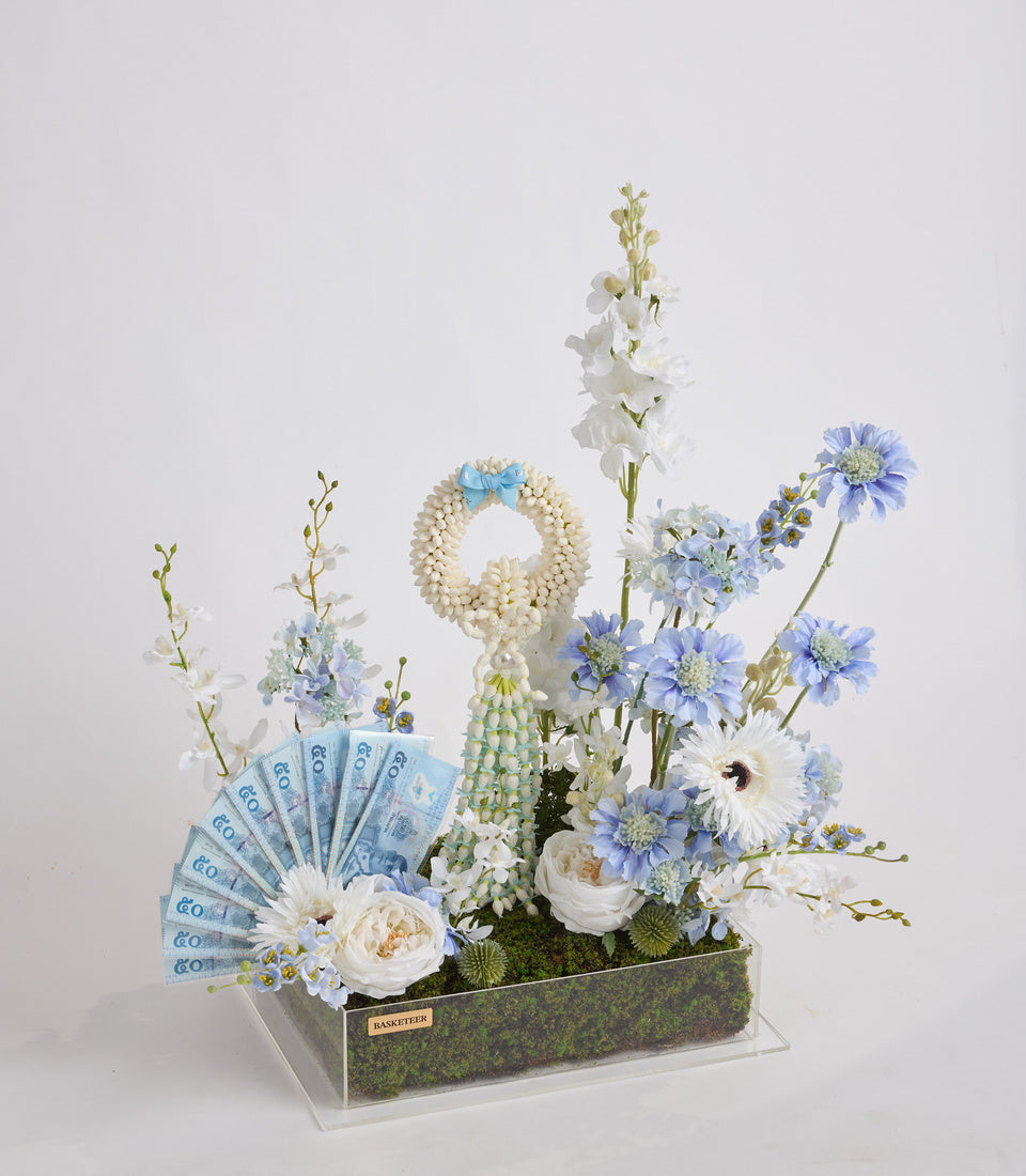 Luxe floral arrangement with white roses, blue daisies, and delicate white flowers in a moss-lined box, accented with blue euro banknotes.