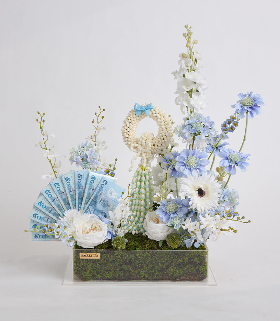Luxury floral arrangement with white roses, blue daisies, pearl beaded peacock sculpture, and fanned euro banknotes in moss-lined container.