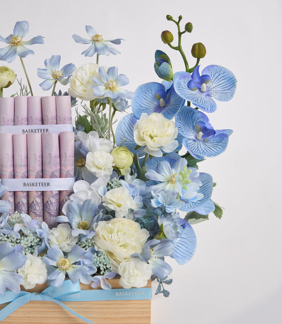 Luxury floral arrangement with blue orchids, white peonies, and cosmos flowers in wooden basket with Basketeer branded packaging and blue ribbon.