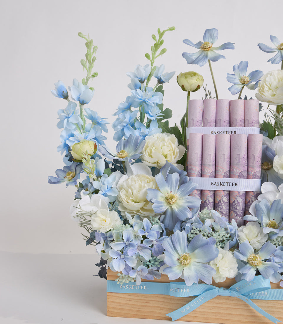 Wooden basket with blue ribbon containing blue delphiniums, white peonies, cosmos flowers, and pink Basketeer product boxes arranged as a luxury gift set.