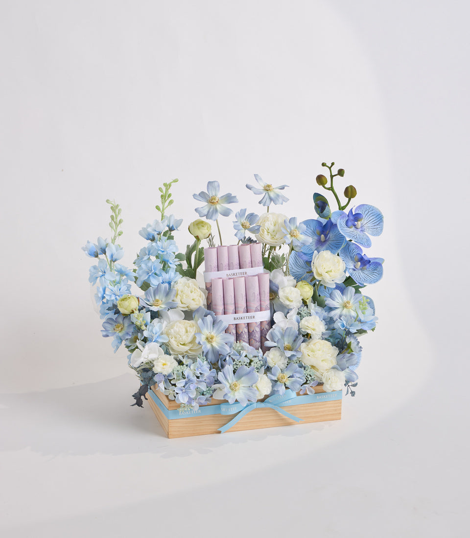 Luxury floral arrangement in wooden box with blue daisies, white roses, blue orchids, and pink BASKET TIER candles, tied with blue ribbon.