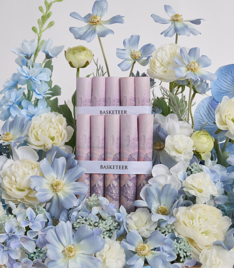 Basketeer luxury candle set with pink cylindrical candles stacked in tiers, surrounded by blue cosmos flowers, white peonies, and delicate greenery on white background.