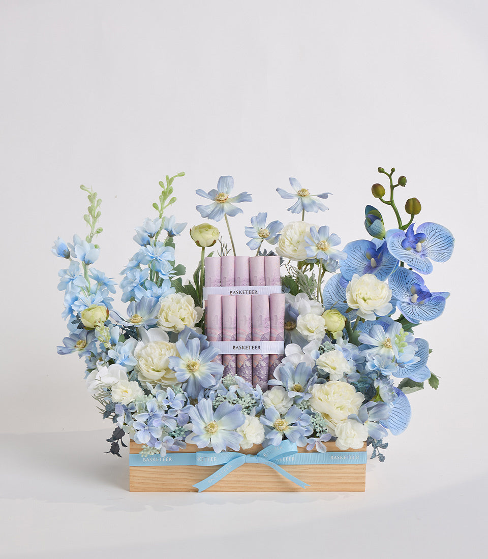 Wooden box with light blue ribbon containing lavender candles surrounded by blue daisies, white peonies, blue orchids, and green foliage.