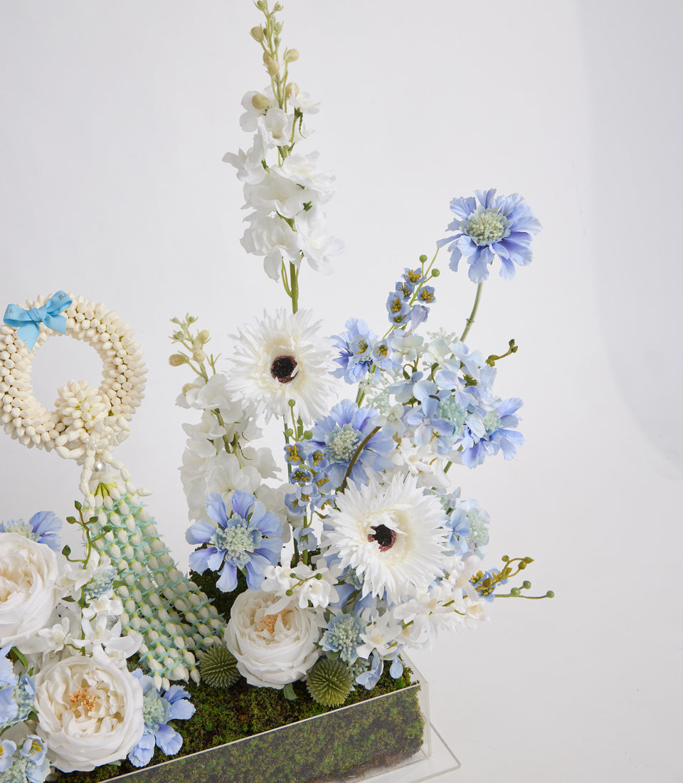 Elegant floral arrangement featuring white roses, blue daisies, white gerberas, delicate white stock flowers, and decorative green pods in a modern rectangular container.