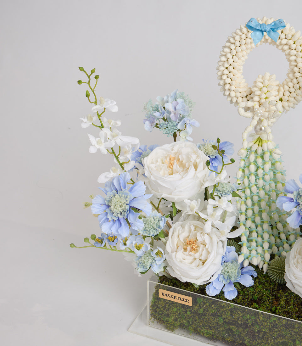 Luxe floral arrangement with cream roses, blue daisies, white orchids, pearl wreath, and moss in acrylic vase with Basketeer label.
