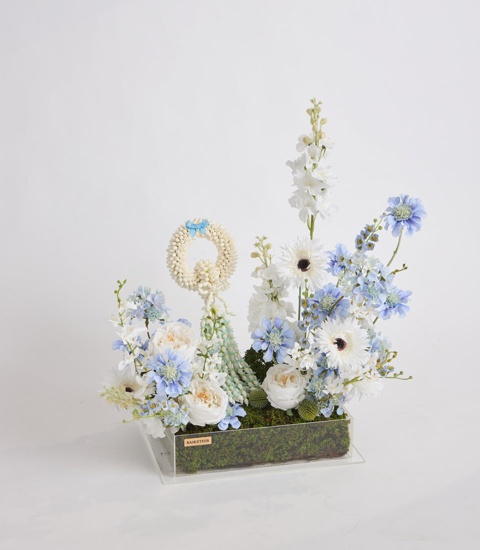 Floral arrangement with white gerberas and roses, blue cornflowers, white delphiniums in clear acrylic box with moss base and gold nameplate.