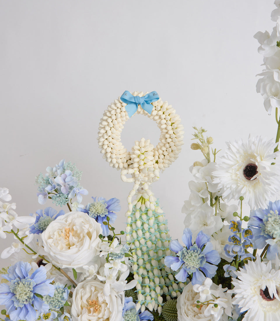 Floral arrangement featuring a cream pearl bead wreath with blue ribbon bow, surrounded by white roses, blue daisies, and white gerberas against a light background.