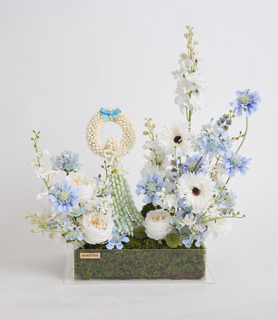 Luxe floral arrangement featuring white gerberas, blue daisies, cream roses, delphinium spikes, a pearl wreath accent, and succulent in moss-lined rectangular container.