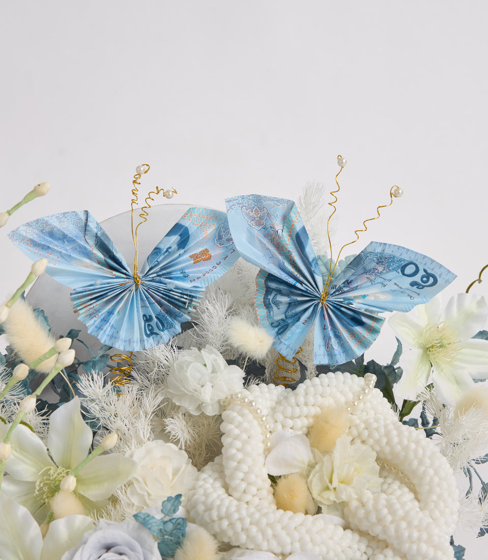 Decorative blue banknote butterflies with gold antennae perched on white flowers, pearls, and silver foliage against a light background.