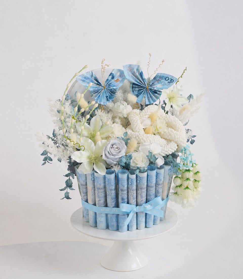 Elegant floral arrangement with white flowers, blue butterflies, and cream-colored rolled papers in a cylindrical vase on a white pedestal stand.