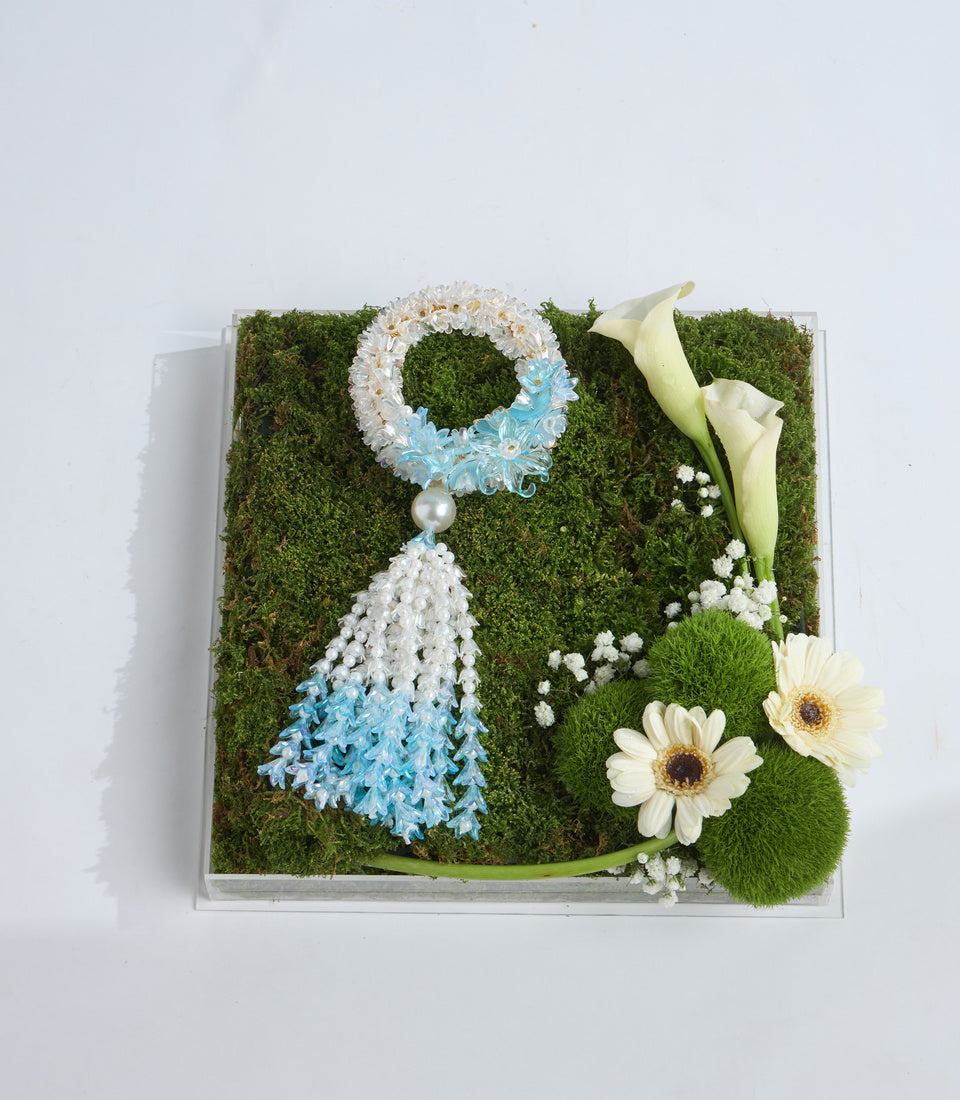 Decorative moss art arrangement featuring white and blue beaded jewelry design, white gerbera daisies, calla lilies, baby's breath, and green moss on white background.