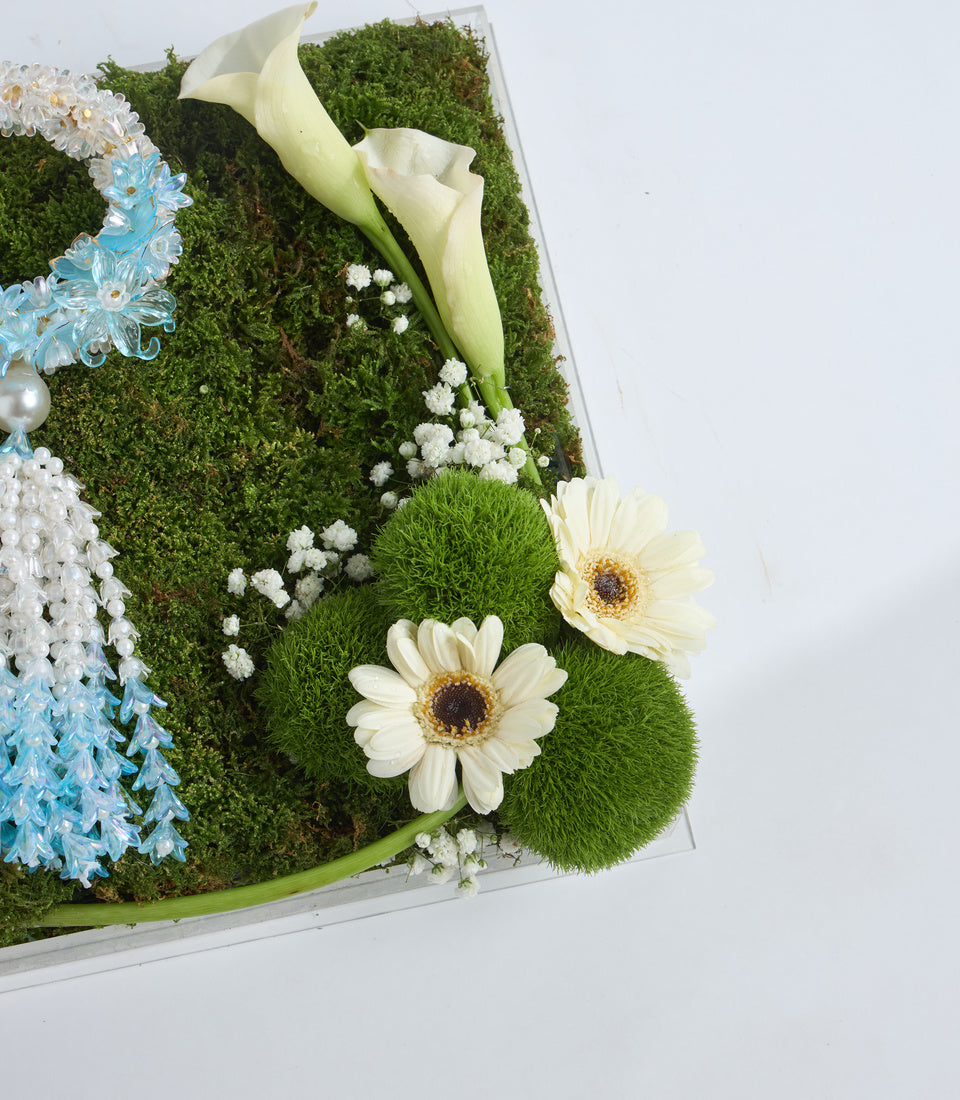 White calla lilies and gerberas with baby's breath on green moss in a rectangular white container, accented with blue flowers and pearl beads.