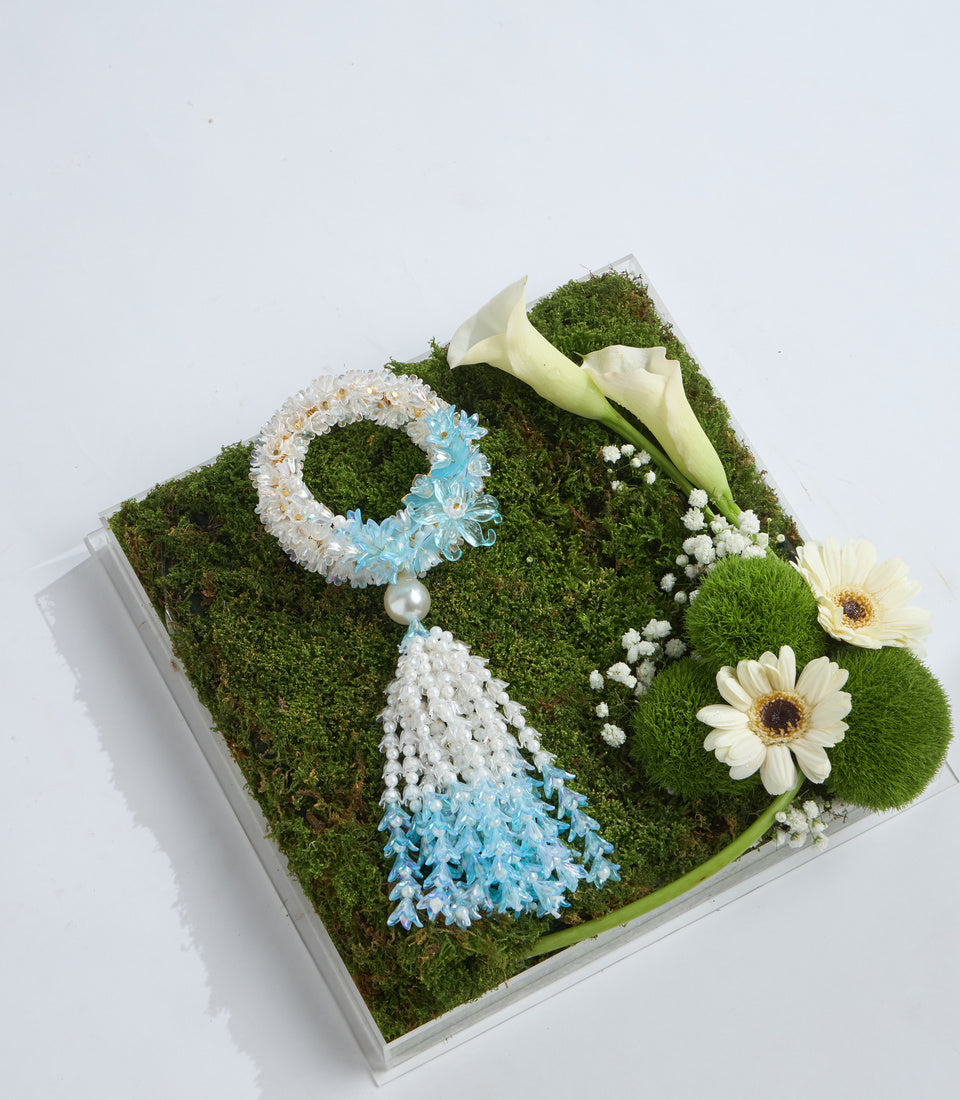 Decorative moss box arrangement featuring white calla lilies, cream gerbera daisies, blue and white flowers, baby's breath, and a pearl accent on lush green moss base.