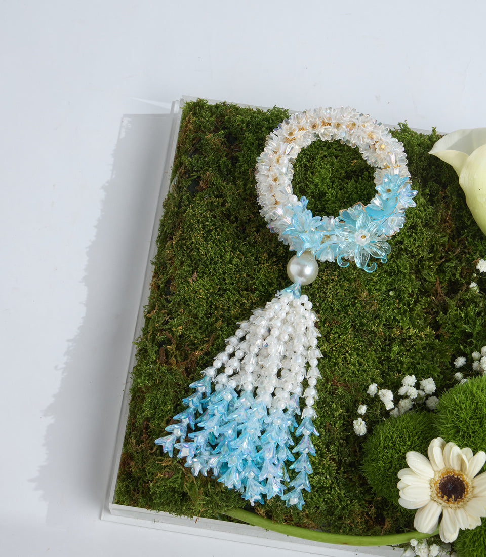 Decorative floral wreath with white and blue daisies, pearl tassel, on preserved moss base in white box.