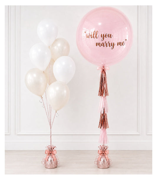 Pink and white balloon arrangement with "will you marry me" text on large pink bubble balloon, rose gold tassels and bows.