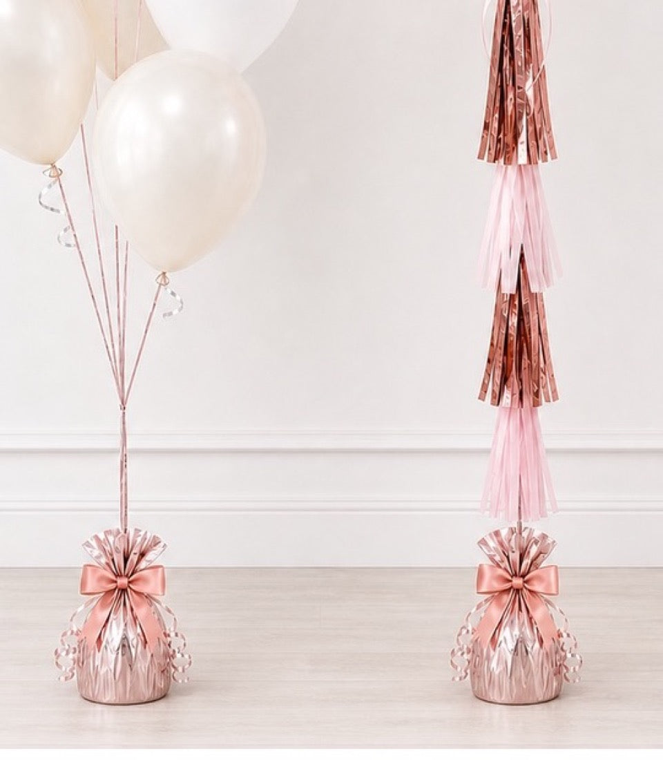 Rose gold balloon weights with pink bows, tassel garlands, and white pearl balloons on wooden floor.