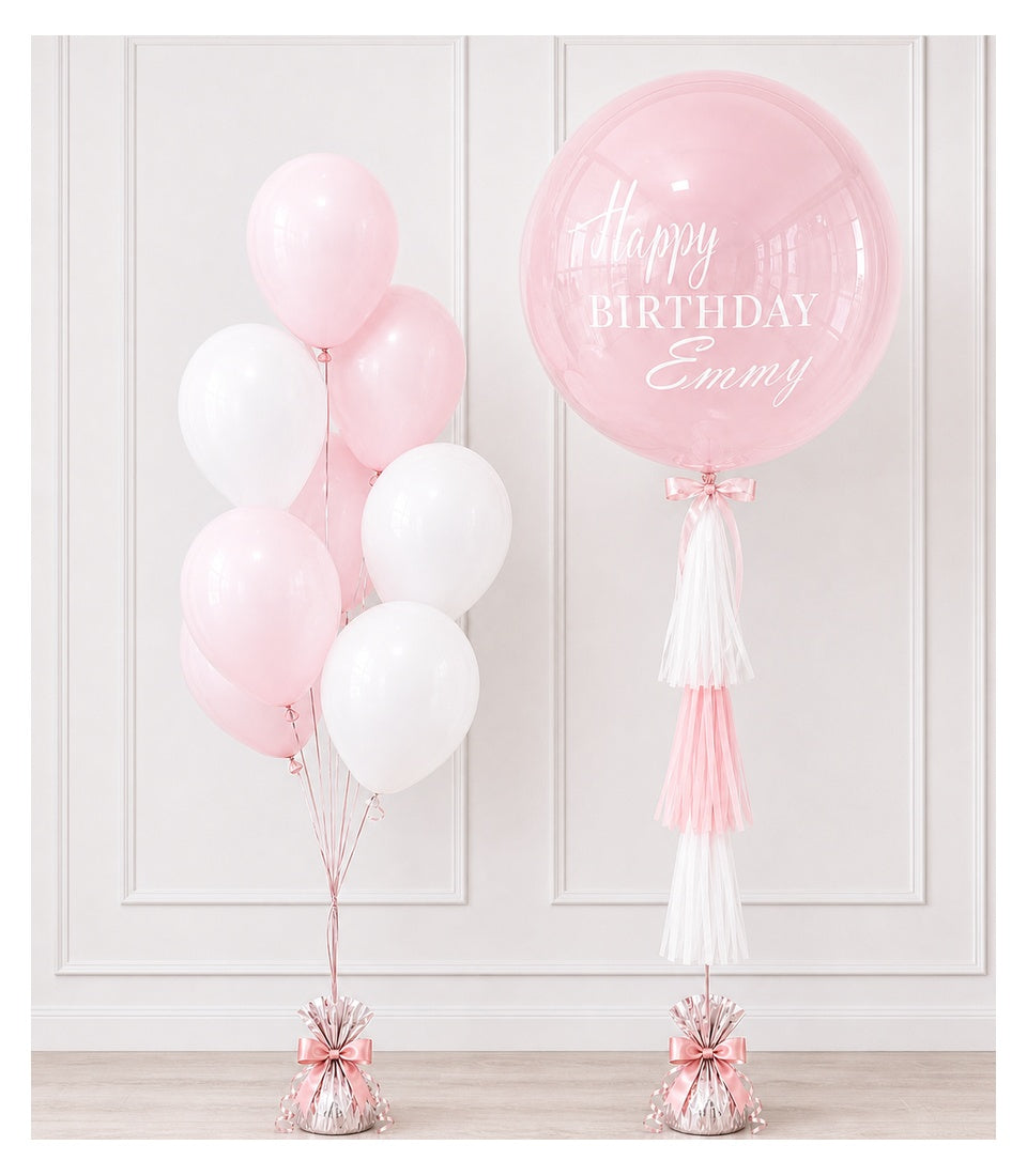 Pink and white balloons with tassels for birthday celebration. Left cluster has six balloons; right giant bubble balloon reads "Happy Birthday Emmy" with pink and white tassel.