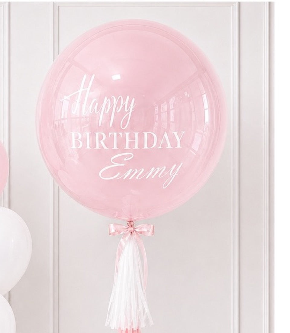 Pink clear bubble balloon with "Happy Birthday Emmy" in white text, decorated with pink bow and white tassel tail.